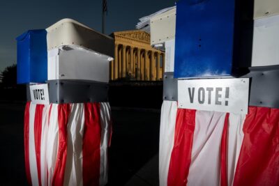 Red white and blue prop ballot boxes in front of a Supreme Court building bathed in crepuscular light.